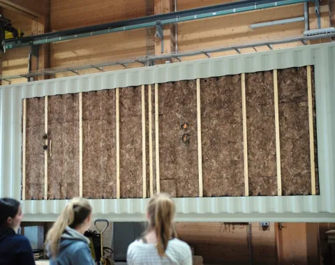 Zwei Personen betrachten eine große, isolierte Wandstruktur in einem Innenraum. Die Wand besteht aus Holz und Dämmmaterial und ist Teil eines modularen Bauprojekts.