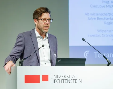 Ein Mann hält einen Vortrag an einem Rednerpult der Universität Liechtenstein. Im Hintergrund ist eine Präsentation zu sehen.