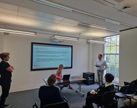 Studierende und Dozenten in einem Seminarraum der Universität Liechtenstein. Eine Präsentation wird auf einem großen Bildschirm gezeigt, während ein Dozent spricht und Studierende aufmerksam zuhören. Helle, moderne Umgebung mit großen Fenstern.