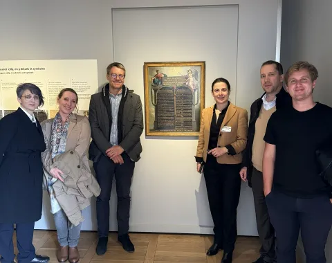 Eine Gruppe von sechs Personen steht in einem Ausstellungsraum vor einem Gemälde. Sie lächeln in die Kamera. Der Raum hat helle Wände und einen Holzboden.