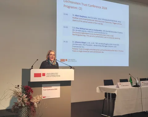Eine Rednerin steht am Podium der Universität Liechtenstein während der Liechtenstein Trust Conference 2024. Im Hintergrund ist eine Präsentationsfolie mit dem Konferenzprogramm zu sehen. Ein Tisch mit Mikrofonen und Wasserflaschen befindet sich rechts im Bild.