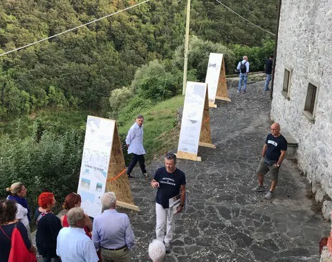 Eine Gruppe von Menschen betrachtet im Freien aufgestellte Tafeln mit Informationen. Die Szene spielt sich auf einem gepflasterten Weg neben einem alten Steingebäude ab, umgeben von üppiger, grüner Landschaft.