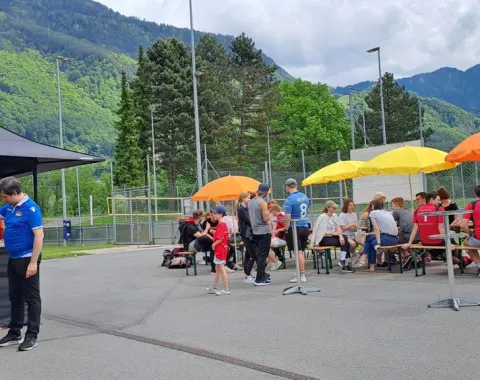 Teilnehmende des Völkerballturniers der Universität Liechtenstein sitzen unter bunten Sonnenschirmen auf Bänken. Im Hintergrund sind Berge und ein Zelt mit Verpflegungsstand zu sehen.