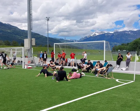 Teilnehmende des Völkerballturniers entspannen sich auf einem Sportplatz vor einer beeindruckenden Bergkulisse. Einige sitzen auf Liegestühlen, während andere am Spielfeldrand stehen. Im Hintergrund sind Fußballtore und Spieler in Sportkleidung zu sehen.