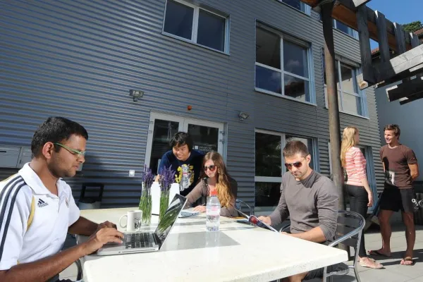 Eine Gruppe von Studierenden der Universität Liechtenstein sitzt und steht um einen Tisch vor der Uni Lichtenstein. Einige benutzen Laptops und Smartphones.