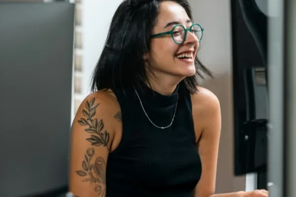 Eine junge lächelnde Frau, mit schulterlangem schwarzen Haar und Brille, in ärmellosem Shirt, das zahlreiche Tattoos entblößt. Sie sitzt vor einem Computer.