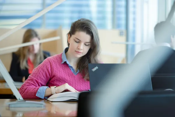 Studentin der Universität Liechtenstein arbeitet konzentriert mit Buch und Laptop in der lichtdurchfluteten Bibliothek.