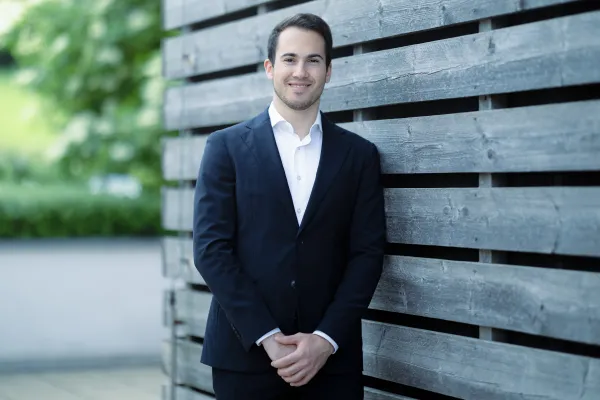 Ein männlicher Studierender im Masterstudiengang Innovative Finance im dunklen Anzug steht entspannt vor einer modernen Holzfassade.