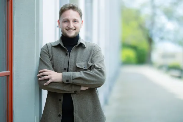 Ein männlicher Studierender im Masterstudiengang Information Systems lehnt mit verschränkten Armen lächelnd an einer hellen Gebäudewand. Er trägt ein braunes Overshirt über einem schwarzen Rollkragenpullover.