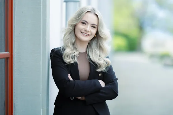 Eine weibliche Studierende im Masterstudiengang Information Systems mit blondem, lockigem Haar, schwarzem Blazer und braunem Oberteil steht an einer Hauswand und lächelt freundlich.