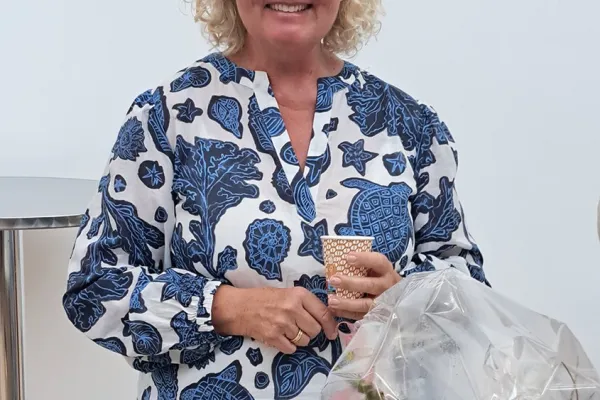 Eine lächelnde Frau mit blonden Haaren und Brille hält einen Becher in der Hand. Sie trägt ein blau-weiß gemustertes Kleid. Vor ihr steht ein Blumenstrauß in Folie verpackt.