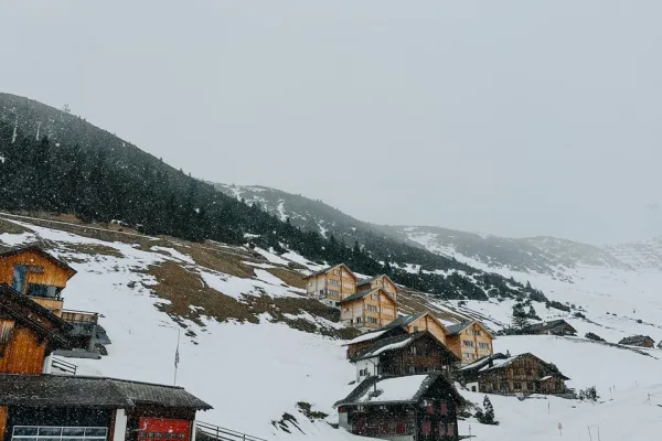 Eine Gruppe von Menschen steht in einer verschneiten Berglandschaft vor traditionellen Holzhäusern. Im Hintergrund sind schneebedeckte Hügel und Nadelbäume zu sehen. Die Szene vermittelt eine winterliche Atmosphäre und zeigt eine Verbindung von Architektur und Natur.