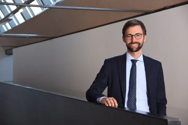 Ein Mann im Anzug steht lächelnd in einem modernen Gebäude mit schrägen Fenstern.