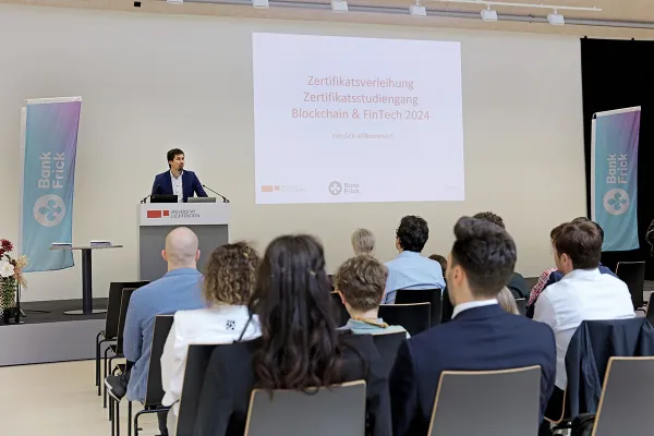 Ein Redner steht an einem Podium vor einer Leinwand mit der Aufschrift "Zertifikatsverleihung Zertifikatsstudiengang Blockchain & FinTech 2024" bei der Universität Liechtenstein. Im Publikum sitzen mehrere Personen. Links und rechts sind Banner von Bank Frick zu sehen.
