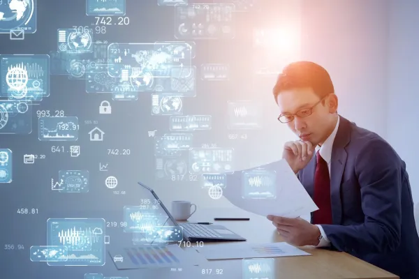 Ein Mann im Anzug sitzt an einem Schreibtisch und betrachtet konzentriert Dokumente. Vor ihm steht ein Laptop, umgeben von digitalen Grafiken und Datenvisualisierungen, die den digitalen Wandel symbolisieren.