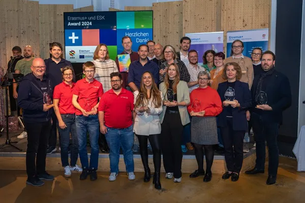Eine Gruppe von Menschen steht auf einer Bühne und hält Trophäen. Im Hintergrund sind Banner mit Erasmus+ Logos und ein Bildschirm mit der Aufschrift "Erasmus+ und ESK Award 2024" zu sehen. Die Personen lächeln und posieren für ein Gruppenfoto.