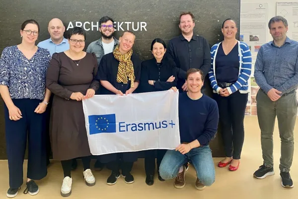 Gruppe von zehn Personen steht vor einer Wand mit dem Schriftzug "Architektur" und hält ein Erasmus+ Banner. Sie lächeln in die Kamera.