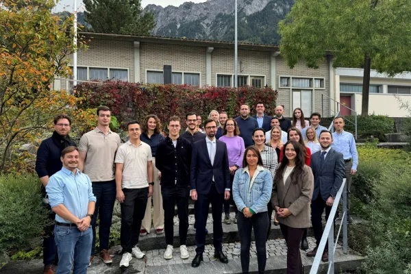 Gruppe von 22 Studierenden und einem Dozenten des Executive Master of Laws (LL.M.) im Bank- und Finanzmarktrecht der Universität Liechtenstein, posierend vor einem Gebäude mit herbstlicher Vegetation und Bergkulisse.