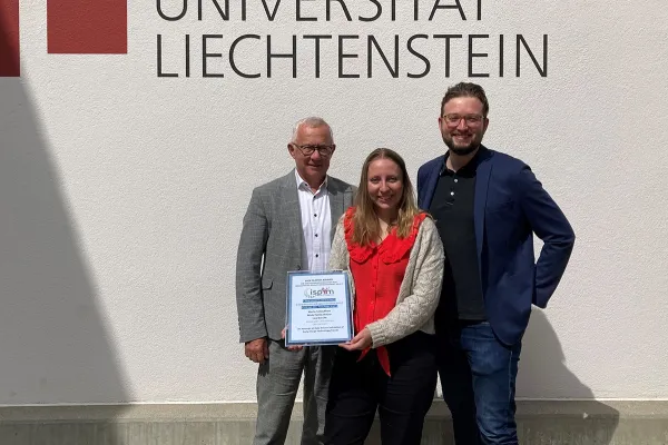 Drei Personen stehen vor einer Wand mit dem Logo der Universität Liechtenstein. Eine Person hält eine Auszeichnung in den Händen.