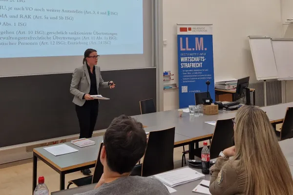 Eine Person hält einen Vortrag in einem Seminarraum der Universität Liechtenstein. Im Hintergrund ist eine Präsentationsfolie zu sehen. Rechts steht ein Roll-Up mit der Aufschrift "LL.M. Wirtschafts-Strafrecht". Teilnehmer sitzen an Tischen und hören zu.