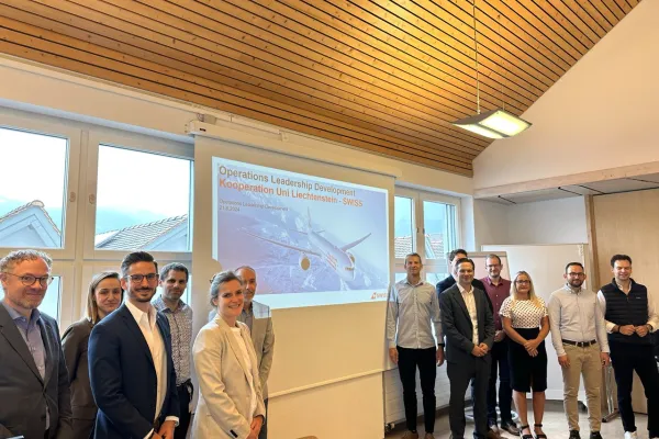 Eine Gruppe von Menschen steht lächelnd in einem Seminarraum vor einer Leinwand, die eine Präsentation zum Operations Leadership Development-Programm der Universität Liechtenstein und SWISS zeigt.