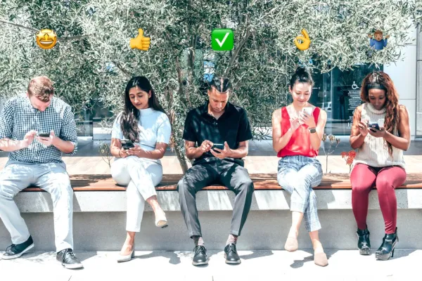 Fünf Personen sitzen auf einer Bank im Freien und schauen auf ihre Smartphones. Über ihren Köpfen sind verschiedene Emojis zu sehen, die Emotionen und Gesten darstellen. Im Hintergrund sind Bäume und ein Gebäude sichtbar.
