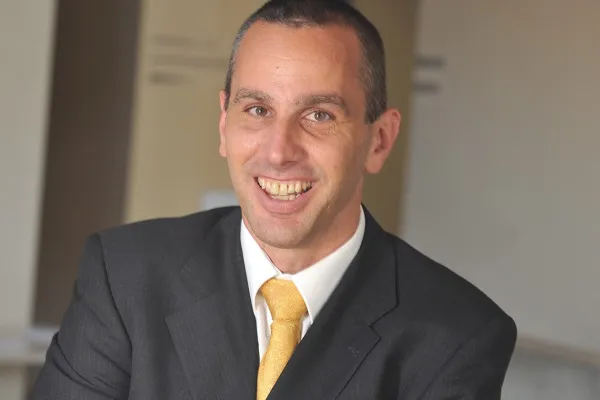 Porträt eines lächelnden Mannes in einem dunklen Anzug mit gelber Krawatte, vor einem neutralen Hintergrund.