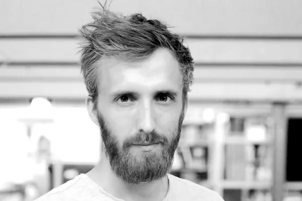 Ein Mann mit Bart und kurzem Haar blickt direkt in die Kamera. Der Hintergrund ist unscharf und zeigt eine Innenraumszene.