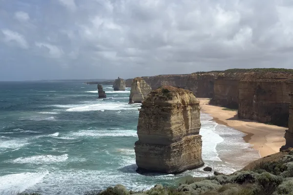 Steile Felsklippen und markante Felsnadeln ragen aus dem Meer entlang einer wilden Küste mit Sandstrand und brechenden Wellen.