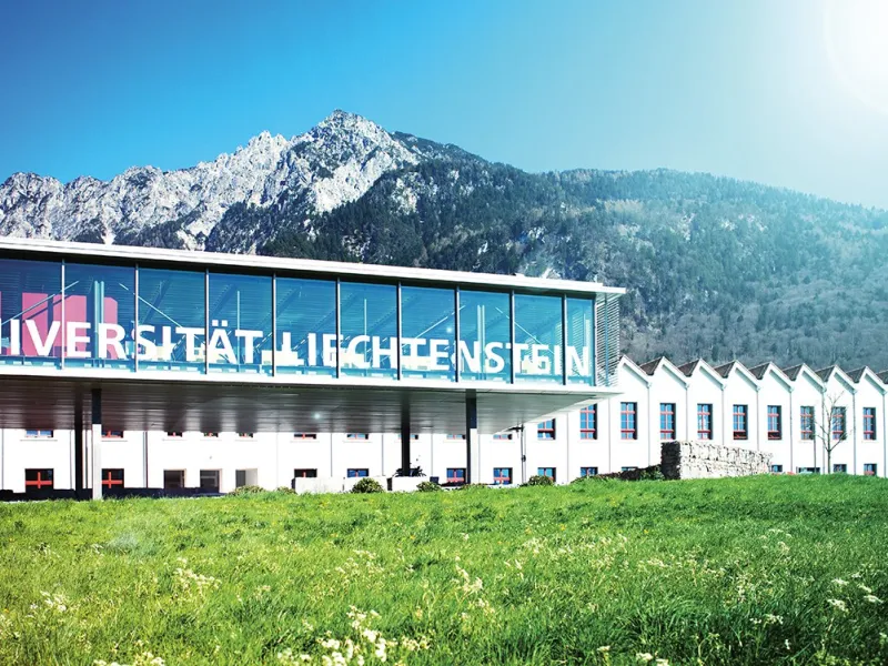 Außenansicht der Universität Liechtenstein mit modernem Glasgebäude und Bergkulisse im Hintergrund, umgeben von grüner Wiese.