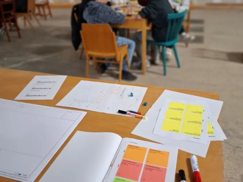 Blick auf einen Schreibtisch mit bunten Notizzetteln und Menschen, die an einem Tisch sitzen im Hintergrund